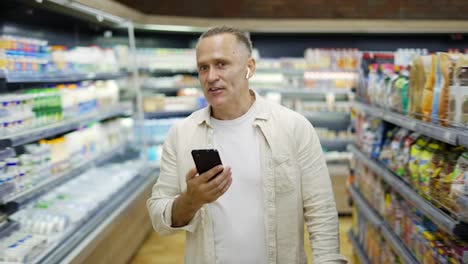 A-middle-aged-man-walks-through-a-grocery-store-talking-on-a-video-link.-Headphones-in-the-ears.-Shopping-trip.-Modern