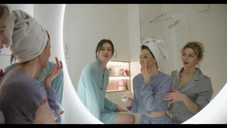 Three-girls-in-bathroom-apply-cream,-talking