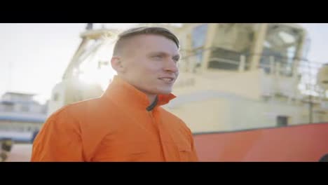 Harbor-master-in-orange-uniform-standing-in-front-of-a-large-ship-in-harbour-and-smiling