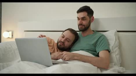 Una-Pareja-Gay-Masculina-Pasa-Tiempo-En-Casa-Tumbada-En-La-Cama,-Usando-Una-Computadora-Portátil