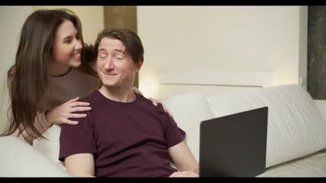 Happy-young-couple-using-laptop-on-sofa,-wife-came-to-hug-her-husband