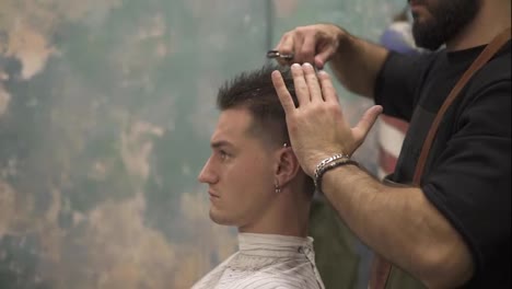Side-view-of-young-handsome-caucasian-man-with-piercing-in-his-ear-getting-his-hair-blow-dried-in-a-retro-stylish-barber-shot