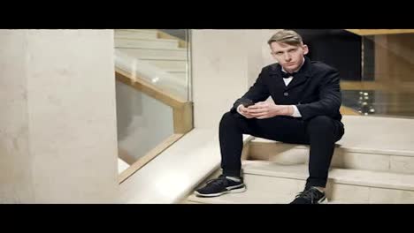Young-blonde-man-in-black-suit-using-his-smartphone-and-sitting-on-stairs-in-hotel.-Waiting-for-someone