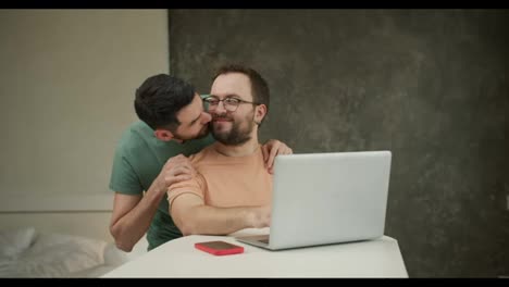 Young-male-hugging-and-kissing-handsome-boyfriend-working-on-laptop