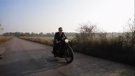 Stylish-cool-young-man-in-sunglasses-and-leather-jacket-riding-motorcycle-on-a-asphalt-road-on-a-sunny-day-in-autumn