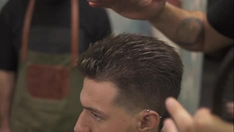 Close-Up-view-of-a-young-handsome-caucasian-man-with-piercing-in-his-ear-getting-his-hair-dressed-and-styled-by-a-bearded-barber