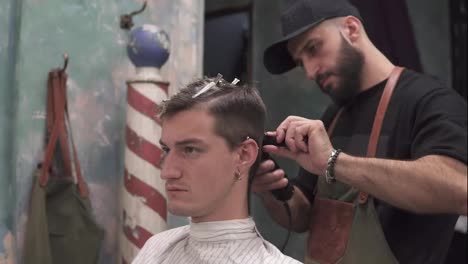 Serious-young-hipster-man-getting-haircut-by-professional-barber-in-barber-shot.-Young-bearded-barber-standing-and-making-stylish