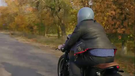 Rückansicht-Eines-Mannes-Mit-Grauem-Helm,-Lederjacke-Und-Kariertem-Hemd,-Der-An-Einem-Sonnigen-Herbsttag-Auf-Einer-Asphaltstraße-Motorrad-Fährt