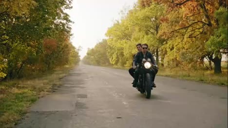 Handsome-man-in-sunglasses-sitting-with-his-girlfriend-behind-the-wheel-of-a-motorcycle-and-riding-on-the-asphalt-road-in-forest