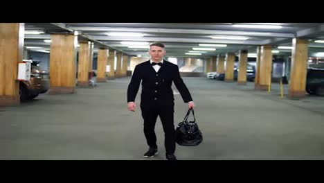 Young-gangster-in-a-black-suit-with-bow-tie-walking-on-the-parking-with-big-leather-bag.-Slow-Motion