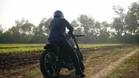 Back-view-of-a-man-in-helmet-and-leather-jacket-coming-up-to-his-bike-and-starting-the-engine-while-standing-on-the-off-road-on-a