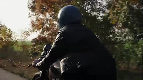 Rückansicht-Eines-Mannes-Mit-Schwarzem-Helm-Und-Lederjacke,-Der-An-Einem-Sonnigen-Herbsttag-Auf-Einer-Asphaltstraße-Motorrad-Fährt.-Bäume-Mit