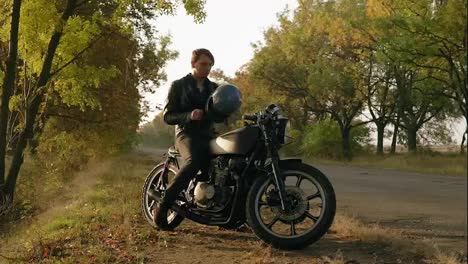 Attractive-man-in-black-leather-jacket-comes-up-to-his-motorcycle,-sits-there-and-puts-a-black-helmet-and-sunglasses-before