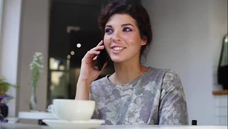 Mujer-Con-Teléfono-Inteligente-Hablando-Por-Teléfono-Mientras-Está-Sentada-En-Una-Cafetería-Con-Una-Taza-De-Café-En-La-Mesa.-Ella-Está-Sonriendo-Y-Riendo-En-El-Café