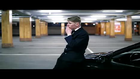 Young-man-in-a-black-suit-with-a-bow-tie-smoking-a-cigarette-sitting-at-the-bonnet-of-the-black-car-in-the-parking