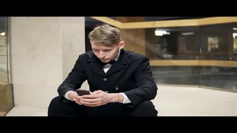 Young-businessman-in-black-suit-with-smartphone-sitting-on-stairs-in-hotel