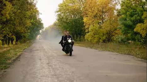 Handsome-man-in-sunglasses-riding-with-his-girlfriend-on-a-motorcycle-on-the-asphalt-road-in-forest-in-autumn.-His-girlfriend-is