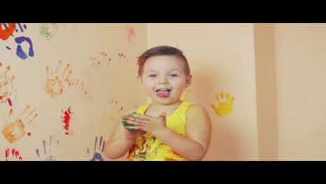 Little-boy-in-yellow-sleeveless-shirt-showing-tongue.-His-hands-are-dirty-in-colors.-He-is-living-his-handprints-on-the-wall