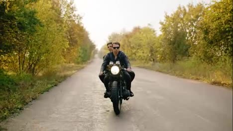 Wide-tracking-shot-of-couple-riding-motorcycle-on-forest-road-in-autumn.-Attractive-young-man-in-sunglasses-driving-his-chopper