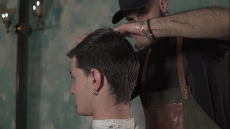 A-hair-stylist-gently-combs-short-damp-hair-of-male-client.-Bearded-barber-in-apron-combing-hair-of-a-male-client-sitting-in-the