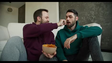 Gay-couple-sitting-on-the-floor-having-a-romantic-conversation-at-home,a-man-feed-his-partner-with-chips