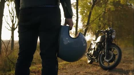 Back-view-of-a-man-in-black-jeans-and-leather-jacket-holding-his-helmet-in-his-hand-while-standing-by-his-cool-bike-on-a-sunny