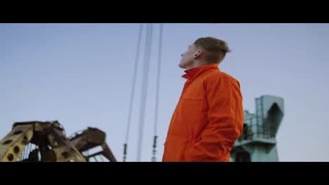 Hübscher-Junger-Containerlagerarbeiter-In-Orangefarbener-Uniform,-Der-Neben-Dem-Schiff-Im-Hafen-Steht-Und-Nach-Oben-Schaut