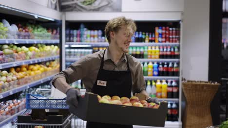 Happy-worker-in-a-black-apron-is-holding-a-crate-of-apples.-Work-in-the-store.-Weekdays.-Healthy-food