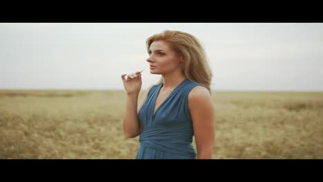 Handsome-young-woman-in-a-long-blue-dress-standing-in-golden-wheat-field-trying-the-wheat's-stem.-Freedom-concept.-Slow-Motion