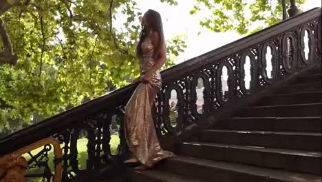Attractive-young-fit-woman-in-a-long-golden-shiny-dress-walks-down-on-the-stairs-on-high-heels-in-summer.-Party-preparation