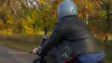 Rückansicht-Eines-Mannes-Mit-Grauem-Helm,-Lederjacke-Und-Kariertem-Hemd,-Der-An-Einem-Sonnigen-Herbsttag-Auf-Einer-Asphaltstraße-Motorrad-Fährt