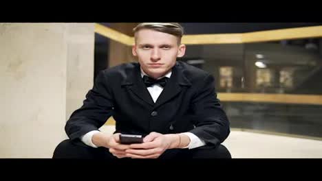 Close-Up-view-of-young-blonde-man-in-black-suit-using-his-smartphone-and-sitting-on-stairs-in-hotel