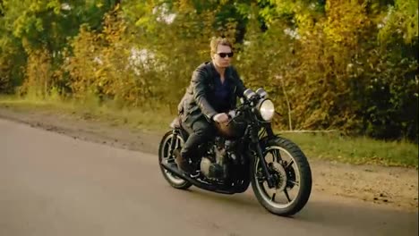 Stylish-man-in-sunglasses-and-leather-jacket-riding-his-cool-bike-on-and-asphalt-road-on-a-sunny-day-in-forest-in-autumn