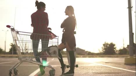 Back-view-of-two-attractive-stylish-girls-standing-by-the-shopping-cart-on-parking-and-talking.-One-girl-in-shorts-is-standing