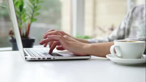 Close-Up-view-of-woman's-hands-working-on-laptop-computer.-Woman-is-sitting-near-the-window-in-cafe-and-typing.-White-cup-with