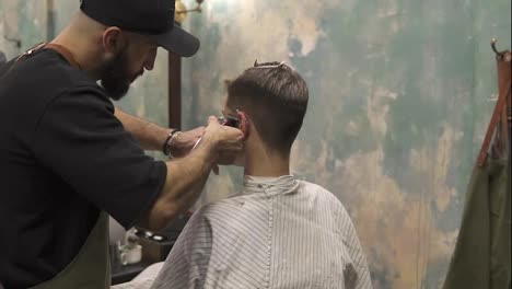 Bearded-barber-creating-a-straight-line-using-electric-trimmer-on-a-man's-temple.-Young-handsome-caucasian-man-getting-a-haircut