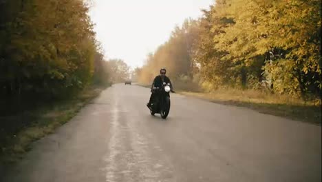 Stylish-young-man-in-sunglasses-and-leather-jacket-riding-motorcycle-on-a-asphalt-road-on-a-sunny-day-in-forest-in-autumn