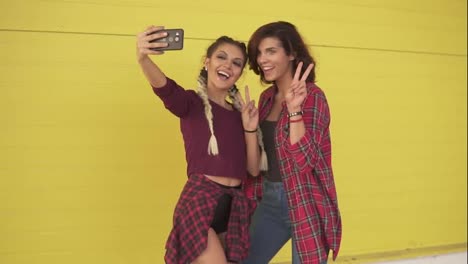 Two-hipster-girls-making-selfie-and-showing-peace-sign-standing-by-the-yellow-wall.-Beautiful-girls-posing-and-taking-photos