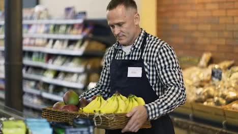 A-middle-aged-saleswoman-in-and-apron-brings-exotic-fruits-in-a-box.-The-concept-of-working-in-a-grocery-store