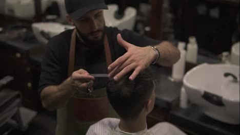 Bearded-barber-in-apron-combing-hair-of-a-male-client-sitting-in-the-chair-at-barber-shot-during-haircut.-Stylish-hairdresser