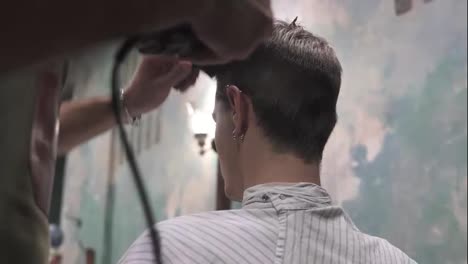 Young-bearded-barber-standing-and-making-stylish-haircut-of-attractive-man-with-clipper-in-barber-shot.-Bearded-man-is-sitting-on