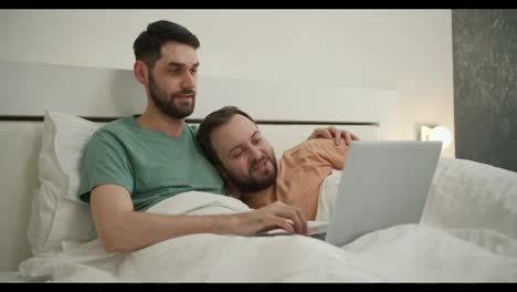 Male-gay-couple-spend-time-at-home-lying-in-bed,-using-laptop-watching-movie