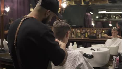 Back-view-of-a-bearded-barber-cutting-hair-using-electric-trimmer-on-the-neck-of-his-customer-in-barber-shot.-Slow-Motion-shot