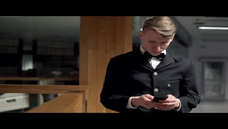 Young-attractive-businessman-standing-near-the-stairs-and-uses-a-smartphone.-He-is-dressed-in-and-expensive-suit-with-a-bow-tie