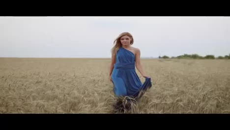 Beautiful-young-happy-girl-in-long-blue-dress-walking-through-golden-wheat-field-looking-to-the-camera.-Freedom-concept