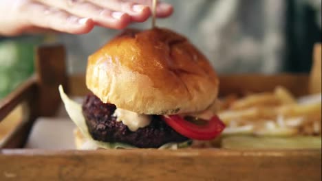 Tasty-burger-with-french-fries-on-a-wooden-tray.-Female-hands-touching-it-to-start-taking.-Slow-Motion-shot