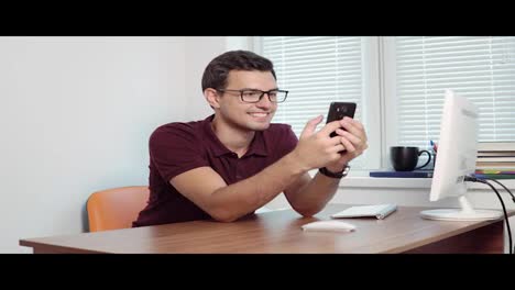 Man-looking-at-the-screen-of-his-phone-and-smiling,-texting-in-the-office-sitting-table.-Handsome-young-businessman-using