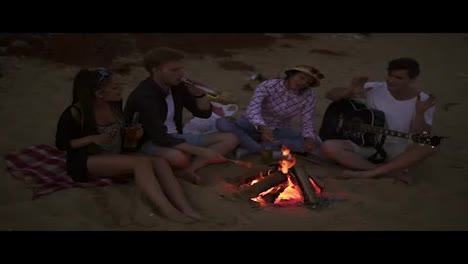 Group-of-young-and-cheerful-people-sitting-by-the-fire-on-the-beach-in-the-evening,-grilling-sausages-and-playing-guitar