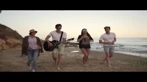 Young-cheerful-happy-friends-walking-on-the-sand-by-the-ocean,-playing-guitar,-singing-songs-and-dancing.-Slow-Motion-shot
