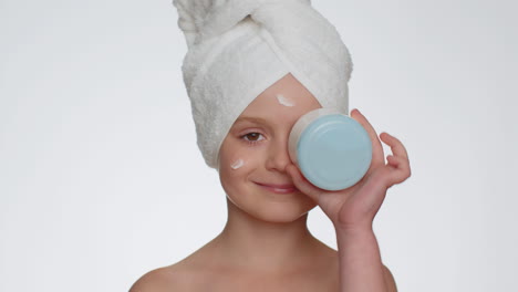 Young-smiling-child-girl-applying-cleansing-moisturizing-cream,-looking-at-camera-holding-creme-jar